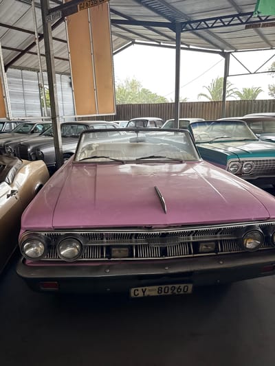 The image shows a collection of vintage cars stored indoors, possibly in a warehouse or garage. The cars are parked close together, with the front of a pink car dominating the foreground. The pink car has a visible license plate that reads "CY 80960". The grills, headlights, and hood of the car are clearly visible. Behind the pink car, other vintage cars in various colors (such as turquoise and silver) are partially visible. The location is suggested to be Kraaifontein, South Africa. The building appears to be a storage facility with corrugated metal walls and ceiling beams. There is a structure of metal beams and supports visible in the lower left. The environment is dimly lit.