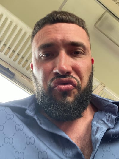 A man with dark hair and a beard is pictured from the chest up, looking into the camera. He has his lips pursed. He is wearing a blue shirt with a pattern of intertwined 'C' shapes. The background appears to be the interior of a vehicle, possibly a train, with off-white ribbed panels and a ceiling vent visible. A sign with Cyrillic text, reading "ВЫХОД" (EXIT), is also visible in the background. The image is taken indoors in what appears to be Izberbash, Russia.