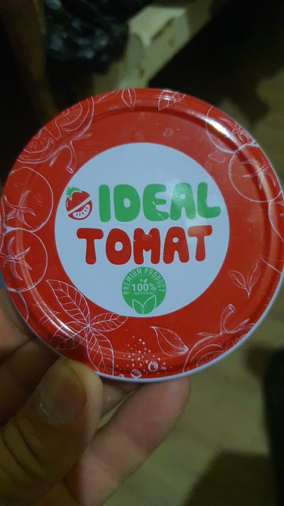 In Karankul, Uzbekistan, a close-up shot captures a red lid of a jar, predominantly featuring a white circular label. The label prominently displays the brand name "IDEAL TOMAT" in bold, block letters. "IDEAL" is in green, while "TOMAT" is in red. To the left of "IDEAL" is a stylized illustration of a tomato with a bite taken out, revealing its seeds. Encircling the bottom portion of the white label is a green circular emblem with the text "PREMIUM PRODUCT 100% NATURAL" and a leaf design. The red lid itself is adorned with white line drawings of tomatoes and leaves. The lid is being held by a person's hand, partially visible at the bottom of the frame. The background is blurred, suggesting an indoor or outdoor setting with muted lighting.