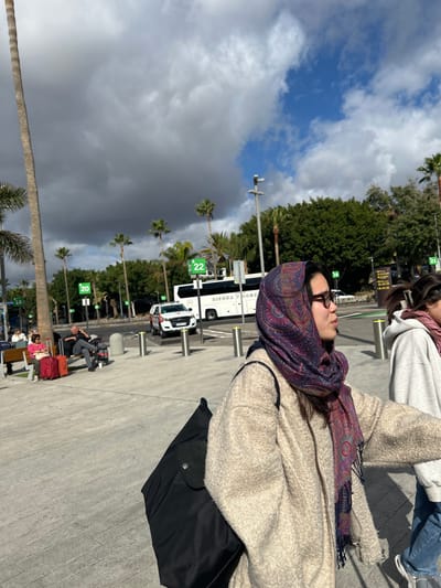 The scene is located in Granadilla de Abona, Spain, at the coordinates 28.048 latitude and -16.577 longitude. It is a bright, sunny day with a few clouds in the sky. A woman wearing a white jacket and a patterned purple scarf is walking toward the camera, a black backpack slung over her shoulder. She is walking past another woman, who is wearing a light grey hoodie and dark pants, heading in the same direction. There is a white van parked in the background. A large white bus with the word "SIERRA" partially visible on the side is further back, behind the woman. Palm trees line the street, and the scene is a typical street in a city.