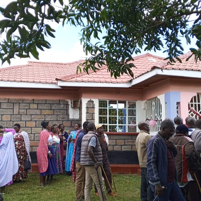 A gathering of individuals, predominantly Kenyan, is situated outside a single-story house in the Keyian ward, Kenya. The house features a red-tiled roof, stone walls, and windows with white trim. Some individuals wear traditional Kenyan clothing. Trees with green leaves partially obscure the view of the house. The ground is covered with green grass. Some individuals are standing, and some have walking sticks. A few balloons are visible near the windows.