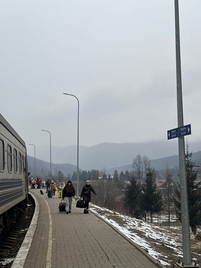 The image, correctly oriented, displays a train station platform scene in Tatariv, Ukraine, under an overcast sky. A group of people, primarily adults, are gathered on the brick-paved platform, appearing to wait for or board a train. Many individuals are dressed in dark, heavy winter coats, suggesting cold weather conditions. They are accompanied by various pieces of luggage, including wheeled suitcases and carry-on bags.
In the foreground, a woman in a dark coat is seen looking towards the viewer with one arm outstretched, possibly waving or gesturing. Further down the platform, other passengers are visible. A train carriage is partially seen on the left, positioned on the tracks adjacent to the platform.
The surroundings include a snow-dusted grassy bank to the right of the platform, with trees, both bare deciduous and evergreens, and distant houses. Rolling hills or mountains are visible in the background, fading into the hazy, grey sky. Several modern streetlights with curved tops are spaced along the platform.
A prominent metallic signpost stands on the platform, displaying two directional signs. The top sign, with an arrow pointing right, reads "ПІВДЕНЬ" and "SOUTH." The bottom sign, with an arrow pointing left, reads "ПІВНІЧ" and "NORTH." The overall impression is of a quiet, chilly day at a rural train stop, characteristic of late winter or early spring.