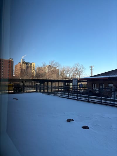 The image shows a winter scene in a city. Clear blue sky above buildings and bare trees. A metal fire escape clings to a brick building. A snowy rooftop dominates the right side of the frame with two dark circular vents. The bottom of the image has a slight reflection of what is being viewed.
