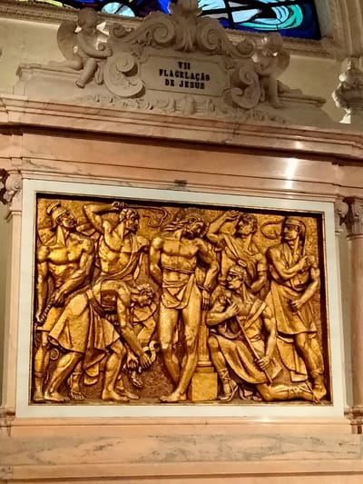 This media file captures a gilded bas-relief sculpture depicting a religious scene, set within a larger architectural context. The central subject is a detailed golden relief featuring numerous figures, framed by what appears to be marble.
The bas-relief portrays the Flagellation of Jesus. In the center, Jesus stands bound to a pillar, looking down in suffering. Around him, several muscular figures, likely Roman soldiers or executioners, are engaged in the act of scourging. One figure on the left raises a whip to strike, while another on the right is also poised for action. Other figures are present, some appearing to prepare tools or observing the scene. The overall composition is dynamic, emphasizing the brutality of the event. The figures are rendered with strong musculature and classical drapery.
Above the bas-relief, an ornate carved marble plaque is visible, flanked by two putti or cherubs. This plaque contains text, identifying the scene. The visible text reads "VII FLAGELLAÇÃO DE JESUS," which translates to "VII Flagellation of Jesus," indicating this is likely one of the Stations of the Cross. In the background, above the marble frame, a glimpse of colored stained glass can be seen, suggesting the setting is a church or religious building.
The entire piece is set against a backdrop of polished, light-colored marble, creating a dignified and solemn atmosphere. The golden material of the relief makes the figures stand out prominently. The image captures a historical religious artwork, likely intended for veneration or instruction within a sacred space like the one found in Fátima, Portugal.