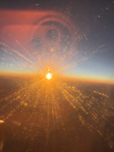 A photograph captures a sunset viewed from an airplane window. The sun, a bright orb of light, is positioned in the center of the frame, emitting golden rays that fan out across the scene. The sky transitions from a deep blue at the top to a soft orange and then a vibrant yellow near the horizon. Below the horizon, the earth is visible as a dimly lit landscape, with hints of city lights appearing as scattered points of light. The airplane window has numerous streaks and smudges, creating a circular, light-refracting effect that overlays the entire image. The scene is bathed in the warm, golden light of the setting sun, creating a dramatic and visually striking composition. The location is Develi, Turkey.