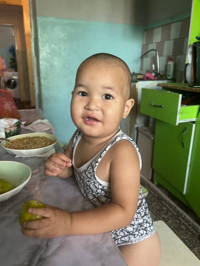 A young child with a shaved head, wearing a black and white patterned tank top, sits at a table in a kitchen. The child is holding a light green fruit in their right hand and looking towards the camera with a slight smile. In front of the child, a bowl of noodles is visible on the table. The kitchen environment includes green cabinets and a sink in the background, with a washing machine and a door visible to the left. The overall scene suggests a domestic setting in the city of Şamaldı-Say, Kyrgyzstan.