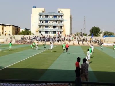 The image shows a soccer match in progress at a stadium in Bahri, Sudan. Players in green and white uniforms compete on a green turf field marked with white lines. A referee in a red shirt and black shorts is visible on the field.

Spectators are gathered in the stands behind the field, some standing along the perimeter. A large banner is displayed along the railing in front of the crowd. In the background is a multi-story building, and other structures are visible in the distance. An electrical transmission tower is also present in the background. Two men stand in the foreground, partially obscured by the fence.