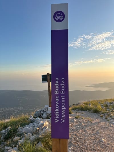 The image shows a viewpoint sign on a mountain in Mirac, Montenegro. The sign is purple with a white outline of binoculars at the top, followed by the text "Vidikovac Budva" and "Viewpoint Budva" in white letters. The sign is mounted on a wooden post. In the background, there is a panoramic view of the sea, islands, and a coastal town, possibly Budva, under a clear sky with scattered clouds. The foreground features a rocky, grassy terrain, typical of a mountain slope. The lighting suggests it is late afternoon or early morning, with the sun casting a warm glow on the landscape.