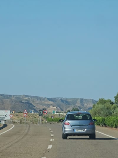 This media file shows a street scene with a silver hatchback car driving on a road. The car is in the foreground, parked on the right side of the road. The road appears to be a winding mountain road with a white line marking the edge. Various traffic signs are visible along the side of the road, including speed limit signs (60 km/h), a roundabout sign, and directional signs indicating "MARIA DE HUERVA," "SANTA FE," "CADRETE," and "zona Industrial." The background features a mountainous landscape with sparse vegetation under a clear blue sky. The time of day appears to be daytime, likely late morning or afternoon, given the lighting and clear weather. The scene suggests a drive through a rural or semi-arid mountainous area, possibly in Spain, given the location context provided. There are no people visible in the image. The car's license plate is visible and reads "1-100 GGM." The overall impression is one of a scenic road trip.