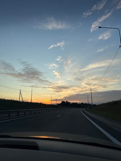 The image depicts a view from inside a vehicle traveling on a paved highway at dusk. The highway curves to the right and is bordered by a metal guardrail on the left and a grassy embankment on the right. Several streetlights are visible along the road, and power lines traverse the sky. The sky is a mix of blue and orange hues, with scattered clouds illuminated by the setting sun. The landscape on either side of the highway is covered in dark vegetation. The front dashboard of the vehicle is visible in the foreground. The specific location is not ascertainable from the visual information.