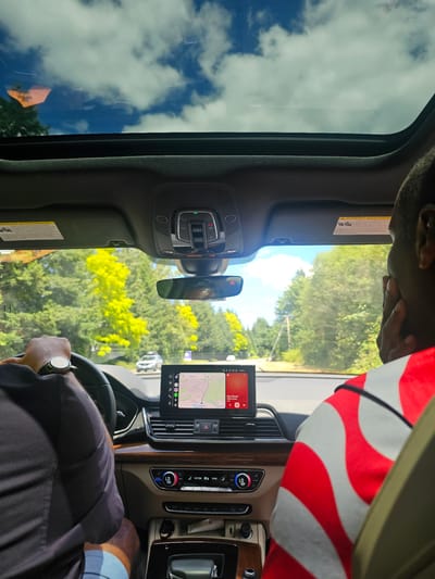 In this image, two individuals are captured inside a vehicle, likely in Issaquah, United States of America, with the camera positioned from the back seat looking forward. The scene is dominated by the interior of a car, with a sunroof revealing a bright blue sky filled with scattered white clouds and lush green trees visible through the windshield. The driver, on the left, is wearing a dark shirt and a watch, and is steering the car. On the right, a passenger is partially visible, wearing a red and white striped shirt and has their hand on their face.
The car's dashboard is prominent, featuring a central infotainment screen displaying a navigation map and music controls. Below the screen are climate control buttons and vents. The overall environment suggests a daytime drive through a suburban or natural area, indicated by the dense foliage and the presence of another car on the road ahead. The lighting is natural and bright, indicating clear weather conditions.