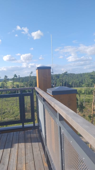 The image shows a view from a wooden observation deck with metal railings. The deck is made of weathered wooden planks and has dark grey metal railings with mesh infills. Two wooden support posts with dark grey caps are visible on the deck, one of which has a tall, thin antenna attached. In the background, there is a scenic landscape of green fields, trees, and a forest under a bright blue sky with scattered white clouds. In the distance, a structure resembling a tower is visible. This scene is located at the Tērvete Nature Park in Tērvete, Latvia.