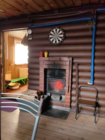 The image depicts the interior of a rustic room, likely a common area or living space within a log cabin. The walls and ceiling are constructed from dark, horizontal wooden logs. In the center of the room, a brick fireplace with a glass-fronted firebox is actively burning, casting a warm glow. Above the fireplace, a dartboard is mounted on the wall. To the left of the fireplace, through an open doorway, a window with bright daylight filtering through illuminates a green bench and a large bowl. Various objects are scattered around the room, including logs near the fireplace, and a metal folding chair positioned to the right of the fireplace. A blue pipe runs vertically along the right wall. The floor is made of light-colored wooden planks. The scene appears to be a domestic, inhabited space, possibly a dacha or vacation home, in the rural setting of Фаніпальскі сельскі Савет, Belarus. There are no people visible in the image.