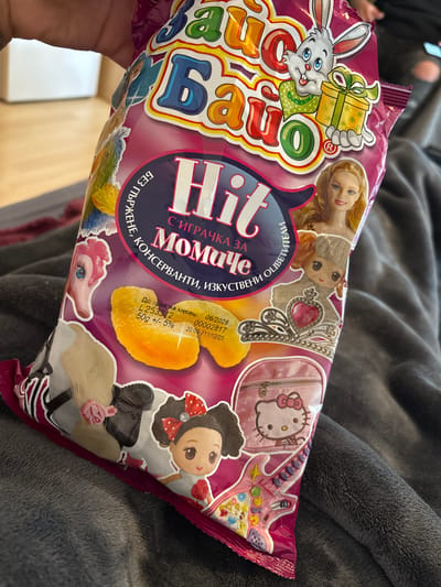 A human hand with a visible tattoo on the wrist holds a brightly colored, magenta-pink snack bag with Cyrillic text and cartoon imagery, positioned over a dark gray, textured fabric surface. The bag prominently displays "Зайко Байо" (Bunny Bayo) at the top and "Hit" centrally, accompanied by the Cyrillic phrase "С ИГРАЧКА ЗА МОМИЧЕ" (With a toy for a girl). Additional text indicates "БЕЗ ГЛЪРЖЕНЕ, КОНСЕРВАНТИ, ИЗКУСТВЕНИ ОЦВЕТИТЕЛИ" (No gluten, preservatives, artificial colors). The packaging features a cartoon white bunny holding a gift, a blonde doll's head resembling Barbie, a cartoon doll with a knitted hat and a tiara, a pink My Little Pony head, a horse's mane with a pink hair clip, a black shoe, a cartoon girl with pigtails and red bows, a pink Hello Kitty backpack, and a pink toy guitar decorated with Disney Princesses. Nutritional information includes a best-before date of 06/2026, a lot number L253312, and a net weight of 50g +/- 5%. The packaging also indicates a production date of 20:09 /11/12/23. The illustration inside the bag depicts several golden-yellow, irregularly shaped snack pieces. The background is out of focus, showing a white wall and light wooden flooring, consistent with an indoor environment. The image contains no discernible visual elements to determine a specific location within Kempten (Allgäu), Germany.