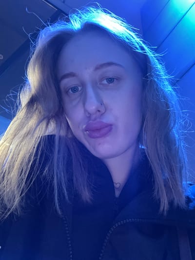 A close-up, rotated image shows a young woman with fair skin and light blonde hair. The lighting is primarily blue, casting a cool tone over the scene. She has blue eyes, a nose piercing on one side, and her lips are slightly parted. A small heart tattoo is visible on her neck. She appears to be wearing a dark jacket or top with a visible zipper and she is indoors. There is no information available about the location, or city Ol'ginskaya, Russia.