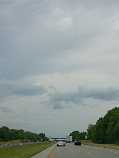 The media file depicts a highway scene in Lexington, United States of America. The perspective is from inside a vehicle traveling on the right lane of a multi-lane highway. The sky is overcast with a mix of light and dark gray clouds, suggesting a cloudy or potentially rainy day. Lush green trees line the right side of the highway, forming a dense border. Orange traffic cones are visible along the shoulder of the road, indicating potential construction or a traffic diversion. Several vehicles are present on the highway, including a white camper van, a white van, and a dark-colored SUV in front of them. Further ahead, there is a sign that reads "DEER CROSSING." The time of day appears to be daytime, given the ambient light. The overall impression is that of a typical highway journey under overcast skies, with some minor traffic and roadwork.