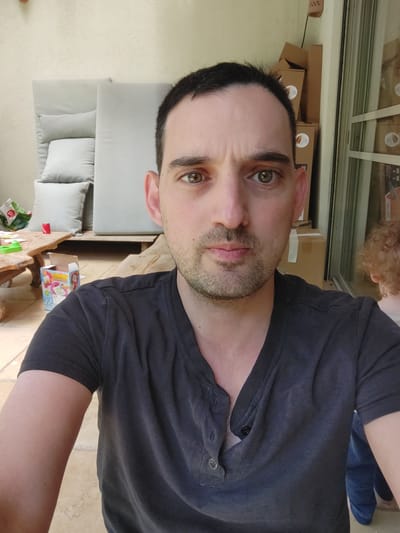 A man is seen from a slightly high, angled perspective, presumably from a selfie taken indoors. He has short dark hair, fair skin, and light brown eyes. He is wearing a dark gray button-down shirt, partially unbuttoned. He has a trimmed beard and mustache. The background includes a beige sofa with pillows, a wooden table with some objects, and several cardboard boxes stacked near a window or glass door. The presence of the boxes suggests a home environment, possibly in Beer Yaaqov, Israel. A child is partially visible towards the bottom of the frame near the window.