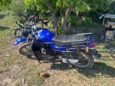 A blue motorcycle is parked on a dirt and grass area under a leafy tree. The motorcycle is facing the right side of the frame and is slightly angled away from the viewer. It has a black seat, black handlebars, and a black rack at the front and rear. The motorcycle has a model number "SL200-K10" visible on its side. In the background, there are more trees, green bushes, and a wooden structure with stacks of lumber. The ground is covered with dry grass, dirt, and some scattered small white flowers. The scene appears to be an outdoor, rural setting. There are no people visible in the image. The location is Oktyabrskoye, Kazakhstan.