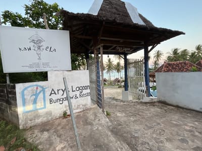 The image shows the entrance to Ary's Lagoon Bungalow & Resto in Legonipah, Indonesia. The entrance is marked by a rustic thatched-roof structure with an open gateway. To the left of the entrance, a white sign displays the name "kaw ela" with a cartoon drawing and a list of offerings: Coffee, Smoothies, Refreshers Drink, Fresh Juice, Dessert, Brunch, and Kitchen. Below this, it states "Open 11 Am - 10 Pm" and a social media handle. Further to the left, a concrete wall features a blue and white logo with the text "Ary's Lagoon Bungalow & Resto". The gateway itself has a white picket fence on the right side and a metal gate on the left, leading to a courtyard with palm trees and bungalows visible in the background. The sky is clear and the overall lighting suggests it is either early morning or late afternoon, likely during a clear weather day. The scene conveys a tropical and relaxed atmosphere typical of a beachfront resort.