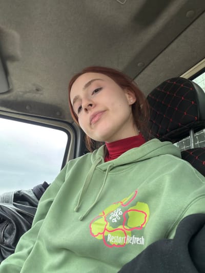 The image displays a single young adult female seated in the passenger seat of a vehicle, located in the city of Siktarva, Georgia. The individual possesses reddish-brown hair and a light complexion, with a small stud earring visible in her left earlobe. She is looking directly at the camera with a neutral expression. Her attire consists of a red turtleneck shirt layered beneath a light green hooded sweatshirt. The sweatshirt features a front graphic depicting a stylized floral design in neon yellow-green with pink outlines, above the text "Restart Refresh" in white lettering with pink outlines. A black garment, possibly a jacket, is partially visible on the left side of the frame. The vehicle's interior shows a light grey ceiling and a black headrest with red diamond-quilted stitching on the right. An overcast or cloudy sky is visible through the vehicle's window on the left, indicating daytime.