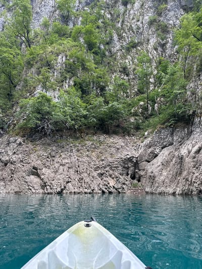 The image shows a serene natural landscape with a rocky cliff face covered in lush green trees. The cliff plunges into the calm, turquoise water, which reflects the surrounding environment. A portion of a white kayak is visible in the lower right corner of the frame, indicating that the photo was taken from the water, likely while on a kayaking excursion. The water's surface is rippled, suggesting a gentle breeze. The overall impression is one of peaceful exploration in a beautiful, unspoiled natural setting. The time of day appears to be daytime, with soft, diffused light suggesting it could be overcast or early morning/late afternoon. The location is identified as Bezuje, Montenegro, which is known for its stunning natural beauty, including canyons and clear waters. There is no visible text in the image.