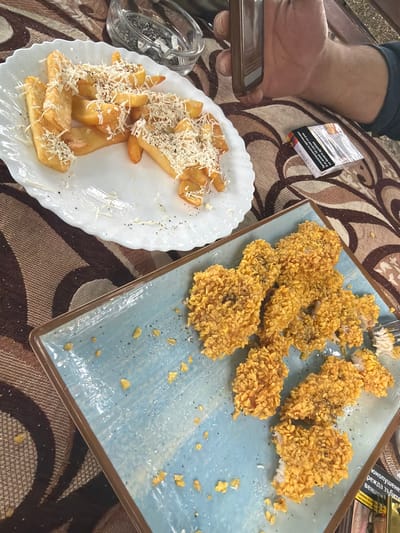 An overhead shot captures a casual dining scene in Sushina, Bulgaria. The image features a table set with two plates of food, a person's hand holding a smartphone, a cigarette pack, and an ashtray, all resting on a patterned tablecloth in brown and beige tones.

On the right, a rectangular light blue plate holds numerous pieces of fried chicken, likely nuggets or tenders, coated in a crunchy, golden, cereal-like breading. Some pieces are sprinkled with black pepper. One piece appears to have been partially eaten, revealing white meat inside. Scattered crumbs of the breading are visible on the plate.

To the left, a white oval plate with a scalloped edge contains thick-cut french fries or potato wedges, generously topped with shredded white cheese and black pepper. Nearby, a glass ashtray holds cigarette butts and ash.

A person's arm and hand are visible on the far left, holding a dark-colored smartphone, suggesting they are either about to eat, taking a photo, or engaging with their device during the meal. A partially obscured pack of cigarettes with Cyrillic text is also on the table.

Visible text on the cigarette pack includes health warnings such as "юнопушенето причинява рак на белите дробове" (smoking causes lung cancer) and mentions "режда зъбите" (damages teeth) and "вени" (gums), reinforcing the Bulgarian context. The bright lighting suggests it is daytime. The overall scene depicts a relaxed meal, possibly at a cafe or restaurant.