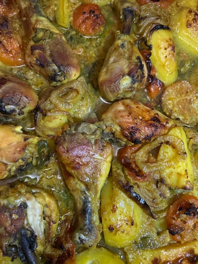 The image shows a close-up of a baked chicken dish. Several chicken drumsticks and thighs are visible, browned and slightly charred in places. The chicken is surrounded by potatoes and sliced onions, all immersed in a light, yellowish sauce. Some sliced tomatoes are interspersed throughout the dish. The overall impression is that of a hearty, homemade meal.