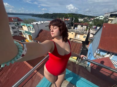 A woman in a red swimsuit is seen from an elevated perspective. She's holding a metal object in one hand and wearing a light blue beaded bracelet on the other. Below her is a cluster of buildings with different colored roofs, followed by a body of water, and a sky filled with clouds. The overall composition is oriented at an odd angle.