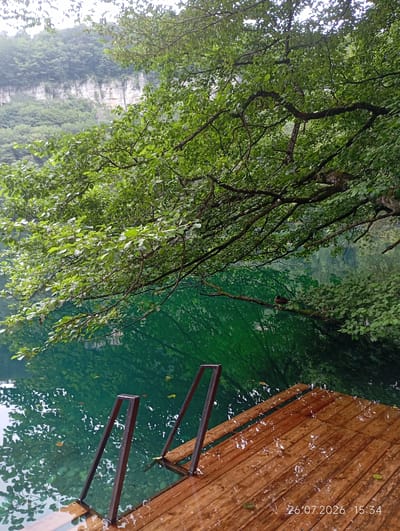 A wooden dock with metal railings extends into a vibrant green lake. The water's surface is partially obscured by reflections of lush green trees and a rocky cliff face in the background. There are two birds perched on a branch overhanging the water. The dock appears wet, with small puddles and streaks visible on the wooden planks, suggesting recent rain. The timestamp "26.07.2026 15:34" is overlaid on the bottom right of the image. The scene is set in a natural environment, likely in the vicinity of Verkhnyaya Balkariya, Russia, given the presence of a lake and mountainous terrain.
