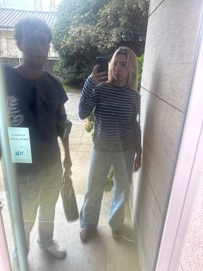 A mirror selfie depicts two individuals, one female and one male, reflected in a glass surface, likely a door, in Cidade da Maia, Portugal. The female, positioned on the right, has blond hair and is wearing a horizontal striped long-sleeve shirt (blue/grey shades), light blue wide-leg jeans, and dark footwear. She holds a dark-colored smartphone in her raised right hand, pointed towards the reflective surface. The male, positioned to the left and slightly behind the female, has dark, curly hair and wears a dark blue t-shirt, light-colored trousers, and white sneakers. He holds a dark bag or object in his right hand, and a dark object is affixed to his upper left arm.
A white rectangular sticker is present on the glass on the lower left, displaying the Portuguese text "POR FAVOR FECHE A PORTA" (Please close the door) and a "jpr" logo. Fine cracks or scratches are visible on the glass surface. The background visible through the glass includes a cobblestone ground, green foliage, trees, and overhead power lines under a bright sky. To the immediate right, a light-grey textured wall and a white door frame are visible. The immediate ground in front of the subjects appears to be light-colored tiles.