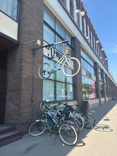A light blue bicycle with a wicker basket is suspended by its front wheel from a metal bracket attached to the brick facade of a building. Below the suspended bicycle, a row of five bicycles are parked on a sidewalk. The building has large windows and is decorated with vertical red and white stripes on its upper section, with several globe-shaped lamps extending from its facade. The sidewalk is paved with concrete, and a shadow of a bicycle is cast on it. The sky is clear and blue. The location is the city of Zhabkino, Russia.