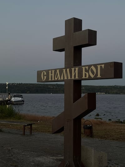 This image depicts a close-up of a large, dark wooden cross positioned by the side of a body of water. The cross is oriented vertically, with its horizontal bar slightly above the center. The letters "С НАМИ БОГ" are inscribed on the vertical beam of the cross in a light, serif font. This translates to "WITH US GOD" in Russian.
The background shows a wide expanse of water, likely a river or lake, with a tree-lined shore on the opposite side. The sky is overcast and gray, suggesting it is either early morning or late evening, or a generally cloudy day. The foreground is a paved walkway with a textured pattern, bordering dry, brown grass. A small, dark brown wooden bench is visible on the walkway near the grass. To the right, a small, dark trash can with a black bag is present. In the water, a modern-looking white boat is docked at a pier, with a few lights illuminated on the pier structure. The overall mood of the image is somber and contemplative, with the cross being the dominant subject. The presence of the water and the overcast sky contribute to a serene, yet somewhat melancholic atmosphere. The location context indicates this scene is in Kazashko, Bulgaria.