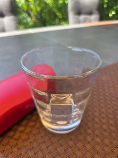 A close-up, eye-level, slightly angled shot shows a clear glass tumbler filled with a clear liquid, resting on a woven brown placemat. To the left of the glass, a bright red, elongated object, possibly a lipstick or a cosmetic item, is partially visible. The background is softly blurred, with the indistinct shapes of greenery and possibly furniture suggesting an outdoor or semi-outdoor setting like a patio or garden. The lighting is natural and somewhat diffused, creating subtle reflections and refractions within the glass. The image was taken in Dissen am Teutoburger Wald, Germany.