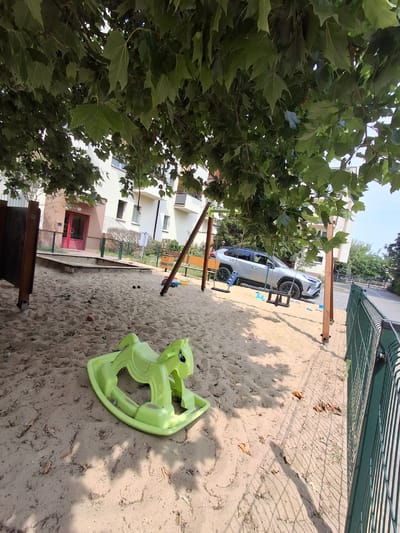 A public playground in the city of Ząbki, Poland, features a sandpit with a light green plastic rocking horse in the foreground. Behind the sandbox, a silver SUV is parked. A swingset with two black swings is visible in the mid-ground, with the wooden frame partially obscured by tree branches overhead. A building with light-colored walls and red accents on the entrance is visible in the background. A green metal fence runs along the right side of the image. The ground is covered in sand and fallen leaves.