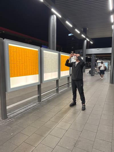 A man wearing a black tracksuit and black sneakers stands on a train platform at night, holding a smartphone to take a picture of a train schedule displayed on a large, illuminated orange board. He is also holding a clear plastic bottle of liquid in his other hand. Another man in a white shirt and dark shorts is walking away from the camera in the background, looking at his phone. The platform is paved with large, grey tiles, and overhead, bright LED lights illuminate the scene. The environment appears to be an outdoor train station with modern architecture, likely in Duisburg, Germany, given the context provided.