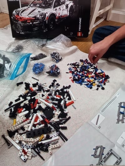 The image depicts an indoor scene, likely a domestic setting, featuring a light-colored, textured rug laid over wooden flooring. The precise location within Brzezina, Poland, is not identifiable from the image. At the rear of the scene, a large black box for the "LEGO TECHNIC Porsche 911 RSR" kit stands upright, displaying an image of the completed model. Several clear plastic bags containing sorted LEGO Technic components, including blue pins and black connectors, are arranged on the rug. Two primary piles of unsorted LEGO elements are present: a larger pile on the lower-left consists of black, white, and red structural beams and various connectors, while a smaller pile on the mid-right contains numerous blue, red, white, and yellow pins and smaller connectors. A person's right arm, clad in dark blue jeans, extends into the frame from the right, with their hand actively selecting pieces from the smaller pile. An open white instruction manual, displaying numbered assembly steps and diagrams, is visible in the lower-right quadrant, indicating an ongoing construction project.