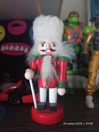 A wooden nutcracker figure with a red coat, white pants, and a white fluffy hat stands in the foreground. The figure has blue eyes, a black mustache, and a white beard. It is holding a white stick with a pinkish ball at the end. Behind the nutcracker, a blurred green Teenage Mutant Ninja Turtle figure is visible. To the right, another blurred figure with dark hair and gold pants stands. The background features a black surface with abstract colorful shapes. A wooden shelf is visible at the bottom, and a red object, possibly a toy car, is on the left side. The scene appears to be indoors, on a shelf. The timestamp "20 июня 2026 г. 19:08" is superimposed on the bottom right corner of the image. The location of Улукаўскі сельскі Савет, Belarus is not visually represented in this image.