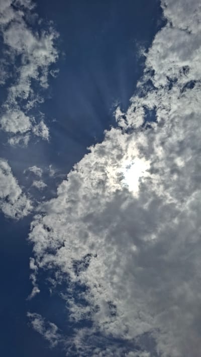 The image shows a sky with scattered clouds. The sun is partially obscured by clouds, creating a bright, hazy effect and casting visible rays of light. The sky is a deep blue, with white and gray clouds of varying densities. There is no indication of human presence or urban features in the image, and therefore no specific location in Gazalkent, Uzbekistan can be identified. The scene is solely focused on the atmospheric conditions.