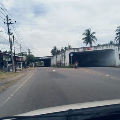 The image depicts a road scene in Cottunong, Indonesia, viewed from inside a vehicle, looking towards a large concrete underpass or tunnel system. The sky is overcast, suggesting a cloudy daytime.
In the foreground, an asphalt road with a white directional arrow painted on it leads towards the underpass. On the left side of the road, multiple utility poles with tangled overhead wires stand beside a row of simple roadside buildings or stalls. While text on these signs is largely illegible due to distance, some appear to feature Indonesian script.
The prominent feature is a wide, weathered concrete underpass structure spanning the width of the road, featuring two large, dark openings. Palm trees grow on top of and behind this structure. Above the left underpass opening, a blue circular sign with a white arrow pointing right is faintly visible. Above the right underpass entrance, a red circular "do not enter" sign is clearly seen. Near this right entrance, a person on a motorcycle is visible, next to a blue circular sign with a white left-pointing arrow. The overall scene suggests a functional, bustling road environment common in semi-urban Indonesian areas.