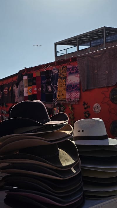 The image captures a close-up view of stacked hats displayed at an outdoor market stall in Puerto Nuevo, Mexico. In the foreground, a tall stack of wide-brimmed hats, predominantly in shades of brown and black, is prominently featured. The hats are arranged in a neat, layered fashion, with some showing signs of wear and creasing. To the right of this stack, a white fedora-style hat with a dark band sits atop another smaller stack of hats.
Behind the hats, the market stall's backdrop consists of a vibrant red wall adorned with various items. Several colorful scarves or textiles with intricate patterns are hanging from a rack. Some of these designs appear to be ethnic or souvenir-like, including one with what looks like a Native American motif and another featuring Hello Kitty. Various t-shirts with logos are also visible hanging on the wall.
Above the stall, a metal structure with a slatted roof suggests an overhead covering or a second story of the market. The sky above is a clear, bright blue, with a single bird in flight visible in the upper left portion of the image. The overall scene suggests a bustling market environment, typical of a coastal town like Puerto Nuevo, known for its lobster restaurants and craft shops. The lighting indicates it is daytime, likely with strong sunlight casting shadows. There are no people directly visible in the frame.