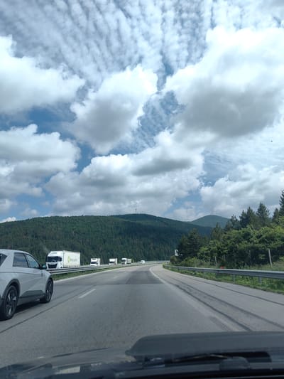 The media file depicts a view from a vehicle traveling on a highway in Potop, Bulgaria. The scene is dominated by a vast, cloudy sky with dramatic formations of cumulus and cirrus clouds against a blue backdrop. Below the sky, a dense, dark green pine forest covers a rolling hillside. The highway itself is asphalt, with clear lane markings and a metal guardrail separating the lanes from the roadside.
Several vehicles are visible on the highway, including a white electric car in the left lane, appearing to be a Hyundai Ioniq 5. Further ahead, a convoy of white trucks, some with trailers displaying logos, are traveling in the right lane. One truck has "Bulog Logistics" visible on its trailer. The time of day appears to be daytime, with bright natural light filtering through the clouds, suggesting partly cloudy to overcast weather conditions. The overall mood is one of transit and natural beauty, with the vast sky and forested landscape creating a sense of openness. The presence of the highway and trucks indicates a scene of travel and commerce.