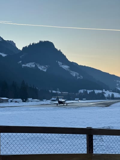 The image captures a winter landscape at Saanen Airport (LSGK) in Saanen, Switzerland. In the immediate foreground, a wooden fence with a wire mesh runs horizontally, partially obscuring a snow-covered field.
A dark, single-engine propeller aircraft, possibly a turboprop, is positioned on a cleared asphalt runway in the midground, facing away from the viewer. Its landing lights are illuminated. The runway is bordered by extensive snow-covered terrain. To the left of the runway, several small, light-colored structures, likely hangars or airport buildings, are visible amidst the snow.
The background is dominated by dark, forested mountains with substantial snow patches on their slopes. Scattered traditional wooden buildings are visible on the lower mountain sides. The sky presents a clear gradient, transitioning from a light blue at the top to a soft orange-yellow near the horizon, suggesting either sunrise or sunset. A distinct white contrail stretches horizontally across the upper sky. No individuals are discernable within the frame.