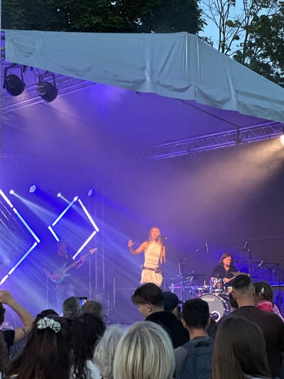 The image depicts an outdoor concert stage at twilight. A female singer, wearing a white, sleeveless top and a white skirt, is performing center stage, illuminated by spotlights. She is holding a microphone and appears to be singing. To her left, a male musician plays a blue bass guitar. To her right, a male drummer is seated behind a drum kit, with the "TAMA" logo visible on the bass drum. The stage is set against a dark backdrop, with elaborate blue and white geometric lighting effects. A large, white canopy extends over the stage, with lighting fixtures attached. In the foreground, a diverse crowd of people are watching the performance, many with their phones raised. The overall atmosphere suggests a lively musical event taking place in Panevėžys, Lithuania.