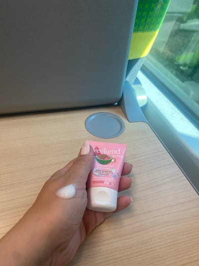 A person's hand, with light-colored fingernails and a subtle shimmer, holds a small, pink tube of "veekend feeling" hand cream. A dollop of white cream is visible on the back of the hand. The hand is positioned over a light wood-grain surface, likely a table. In the background, a grey textured surface and a window with a view of green foliage are visible. The scene appears to be inside a train compartment, given the seating arrangement and window design. The location is not Sopron, Hungary.