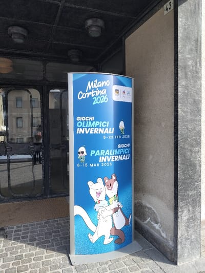 A blue, curved vertical banner promoting the Milano Cortina 2026 Winter Olympic and Paralympic Games stands on a cobblestone sidewalk in Serdes, Italy. The banner displays "Milano Cortina 2026" at the top, followed by "GIOCHI OLIMPICI INVERNALI 6-22 FEB 2026" and "GIOCHI PARALIMPICI INVERNALI 6-15 MAR 2026." Cartoon mascots, including two stoats hugging at the bottom and two smaller figures near the game titles, are visible on the banner.

The banner is positioned to the left of a building entrance, which features double glass doors with dark, ornate metal frames. A dark, textured canopy with three recessed light fixtures extends over the entrance. Inside the doorway, a brown doormat is visible. Faint reflections of a person in a light-colored top and other buildings are discernible in the glass doors. To the right of the banner, a light brown concrete or stone building facade displays the dark house number '43'. The scene is illuminated by direct sunlight, casting shadows on the cobblestone ground to the left of the banner.