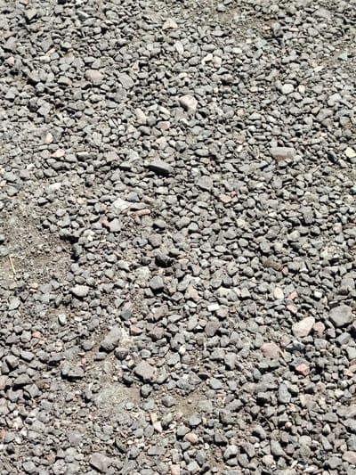 The image displays a close-up, overhead view of a surface covered in a dense layer of small, irregularly shaped stones and gravel. The stones vary in color, predominantly shades of gray, with some lighter tan and pinkish hues interspersed. They are closely packed, creating a rough and uneven texture. Small amounts of dark soil or dust are visible in the crevices between the stones. The lighting suggests an outdoor environment with shadows cast by the stones, indicating sunlight. There are no discernible people, animals, or structures in the image. The scene appears to be a common ground surface, such as a pathway, driveway, or unpaved area. The location is identified as being in the city of Torangalyk, Kazakhstan.