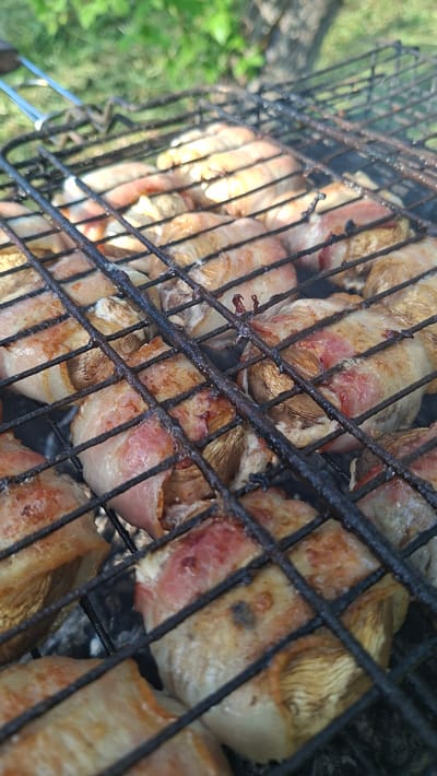 The image shows mushrooms wrapped in bacon grilling on a black metal grill. The grill appears to be outdoors, as evidenced by the green vegetation visible in the background. The focus is on the food, with the grill and background slightly blurred. There's some smoke present. The mushrooms are tan and appear mostly cooked, with some white parts still visible. The bacon is pink and appears to be sizzling. The image seems to be taken from a slightly elevated angle. The location is likely Сваткаўскі сельскі Савет, Belarus.