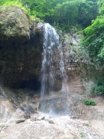 The image shows a waterfall cascading down a rocky cliff face, surrounded by lush green foliage. The waterfall is the primary focus, with water fanning out as it falls. The cliff is composed of layered rock, with patches of moss and small plants growing on it. To the left of the waterfall, a large, moss-covered rock overhang forms a natural alcove behind the falling water. The ground in the foreground is covered in small rocks and pebbles, with a small stream of water visible at the bottom. The environment suggests a natural, outdoor setting. The location is identified as Khushto-Syrt, Russia. There are no people or animals visible in the image.