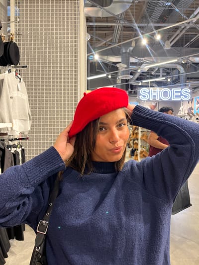 A young adult female is depicted from the chest up, centered within a Primark retail store located at 1621 Chestnut St, Philadelphia, PA 19103. She wears a dark blue, textured long-sleeved sweater and a black shoulder bag with a silver buckle. She holds a bright red beret on her head with both hands. Her lips are pursed, and she gazes directly at the camera. The background to her left features a white grid partition and clothing racks displaying garments, including a grey sweatshirt with "SAN DIEGO" printed in white. To her right, a blue neon sign displays "SHOES" in uppercase letters. A partially obscured individual is visible behind her right shoulder. The overhead environment consists of exposed silver ductwork and multiple industrial light fixtures.