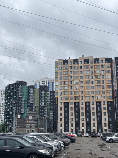 The image shows a row of modern, multi-story buildings and a parking lot filled with cars. The buildings feature a mix of dark and light-colored facades, with numerous windows and balconies. Some of the buildings have a distinctive pattern of alternating dark and light square blocks. One building has a facade with numerous rectangular windows framed in wood-like material. Another section of buildings displays a color scheme of green and black.
In the foreground, a parking lot is visible with many cars parked in a line. The cars are of various colors and models, suggesting a busy urban or residential area. The ground of the parking lot appears to be paved with asphalt or similar material and shows some signs of wear or discoloration.
In the background, the tops of the buildings are visible against a cloudy sky. Several power lines run horizontally across the image, suggesting an overhead electrical system. One of the buildings has visible signage that appears to be in Cyrillic script. A large clock face is also visible on one of the buildings, indicating a daytime setting. The overall impression is of a residential or mixed-use development in an urban environment. The weather appears to be overcast, suggesting a cloudy day. There are no people clearly visible in the scene. The location is identified as Ligachevo, Russia.